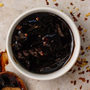 A small white bowl filled with thick, glossy spicy balsamic glaze topped with red pepper flakes, surrounded by scattered chili flakes on the counter.