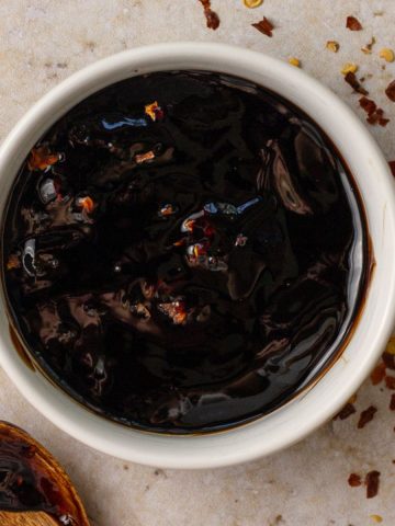 A small white bowl filled with thick, glossy spicy balsamic glaze topped with red pepper flakes, surrounded by scattered chili flakes on the counter.