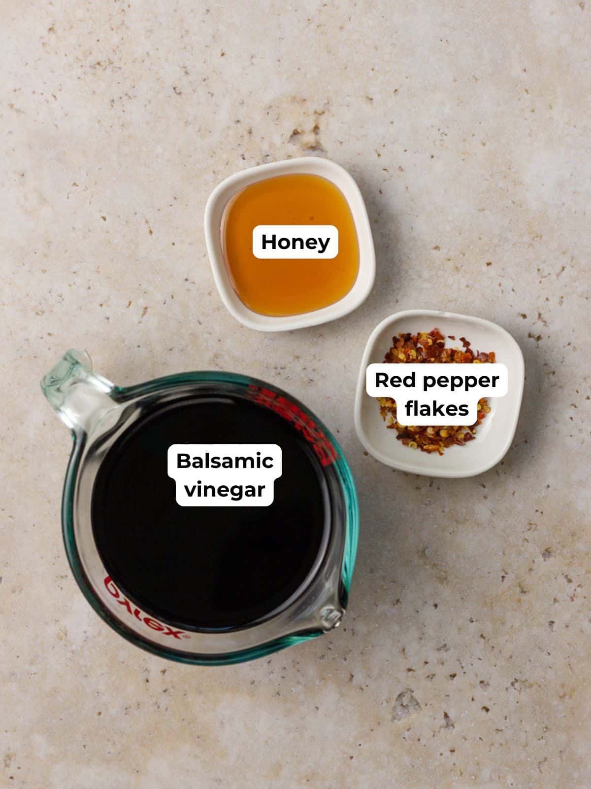 A measuring cup of balsamic vinegar sits next to small bowls of honey and red pepper flakes on a neutral countertop.