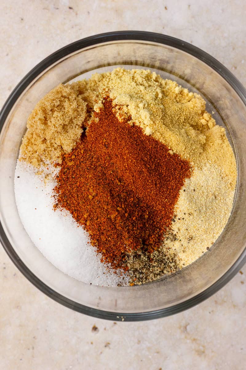 A bowl of unmixed sweet and smoky rub ingredients showing colorful layers of spices and brown sugar.