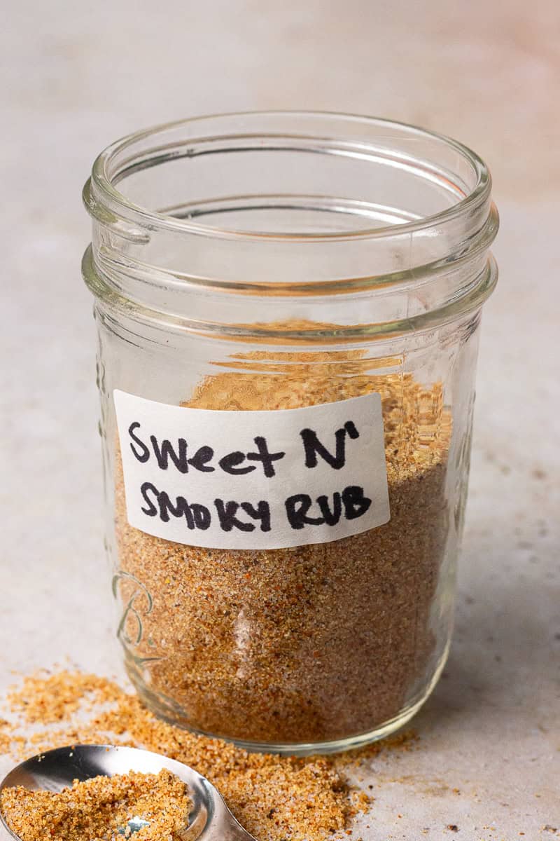 A labeled mason jar of sweet and smoky rub with some spilled seasoning and a measuring spoon beside it.