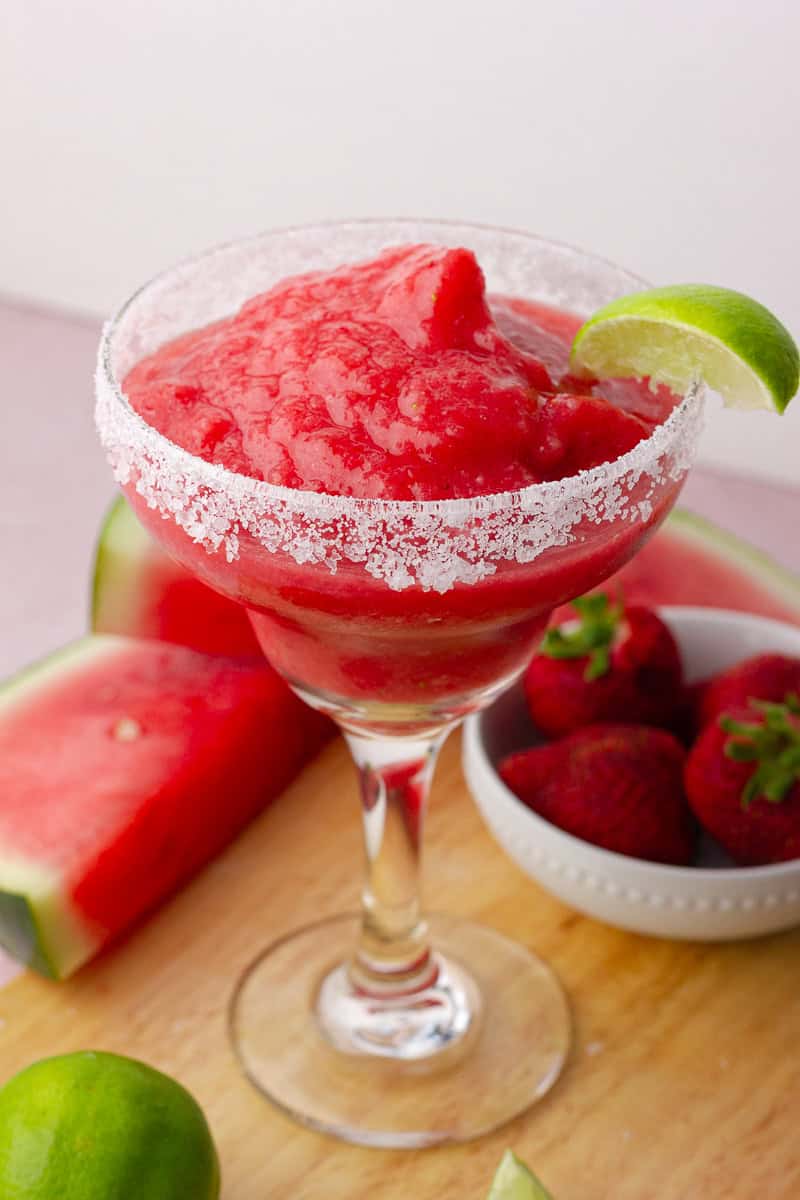 A frozen strawberry watermelon margarita in a salt-rimmed glass with a lime wedge, surrounded by fresh fruit.