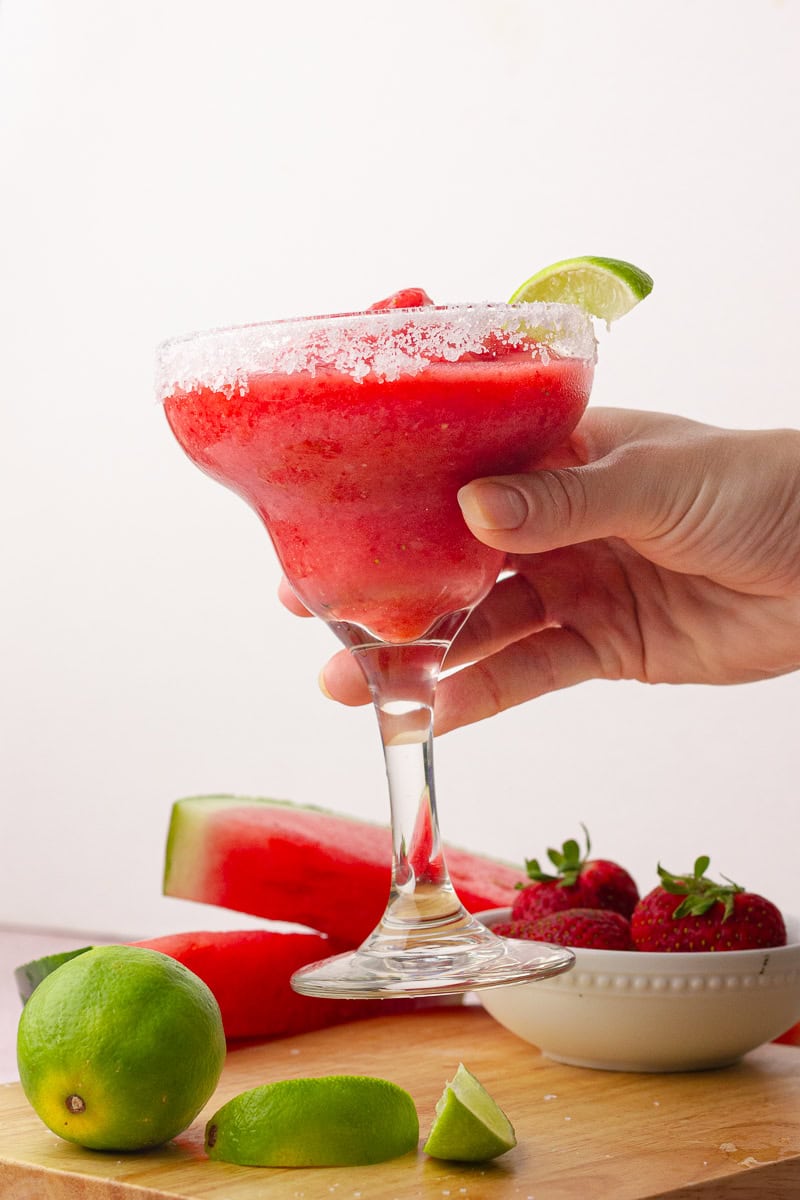 Hand holding a salt-rimmed margarita glass filled with frozen strawberry watermelon margarita over a table with fresh fruit.