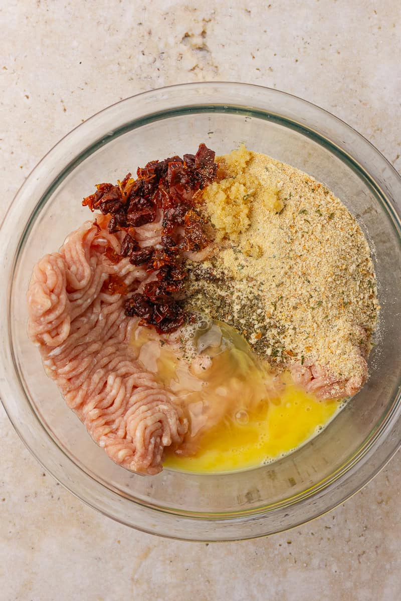 A mixing bowl filled with ground chicken, breadcrumbs, chopped sun-dried tomatoes, minced garlic, and a cracked egg ready to be combined for burger patties.