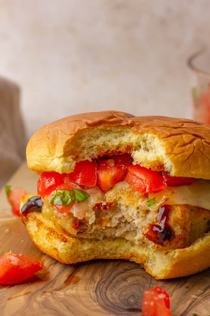 A juicy chicken burger on a bun with a bite taken out, topped with fresh tomatoes, basil, and melted provolone cheese.