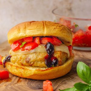 A golden, juicy bruschetta chicken burger topped with melted provolone, fresh diced tomatoes, basil, and a drizzle of balsamic glaze on a toasted bun.