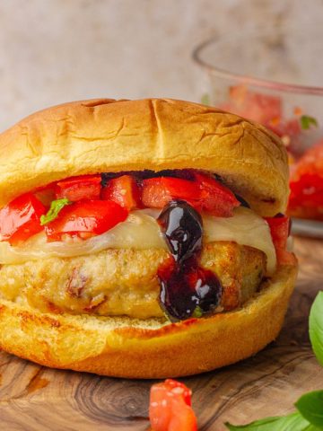 A golden, juicy bruschetta chicken burger topped with melted provolone, fresh diced tomatoes, basil, and a drizzle of balsamic glaze on a toasted bun.