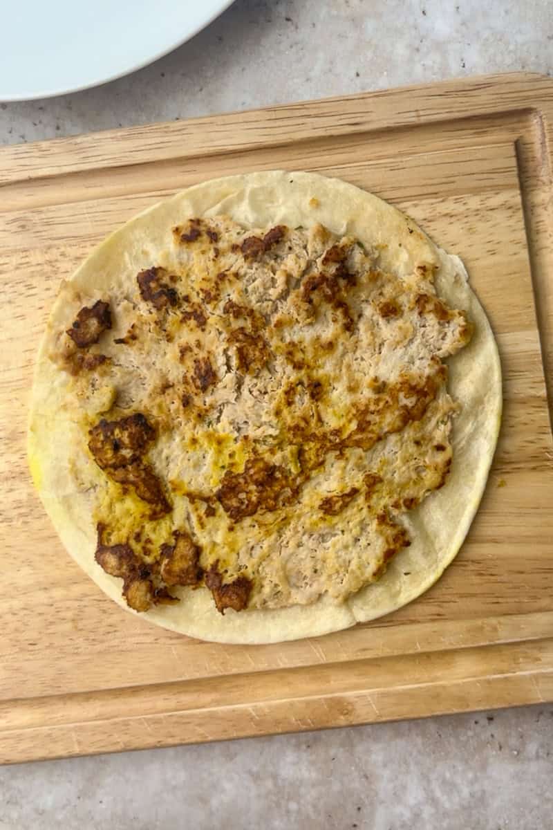 Smashed ground chicken cooked on a flour tortilla sits on a cutting board, ready for toppings.
