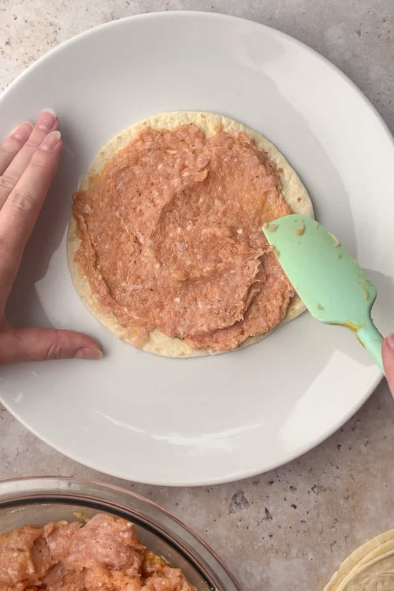 A silicone spatula spreads seasoned ground chicken in a thin, even layer on a flour tortilla.
