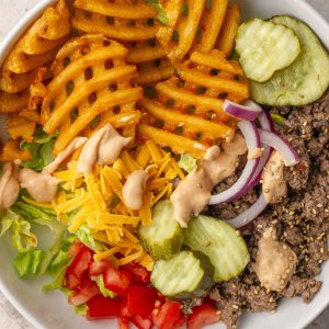 A loaded burger bowl with waffle fries, ground beef, pickles, tomatoes, red onion, cheddar cheese, and special sauce.
