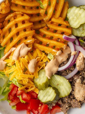 A loaded burger bowl with waffle fries, ground beef, pickles, tomatoes, red onion, cheddar cheese, and special sauce.