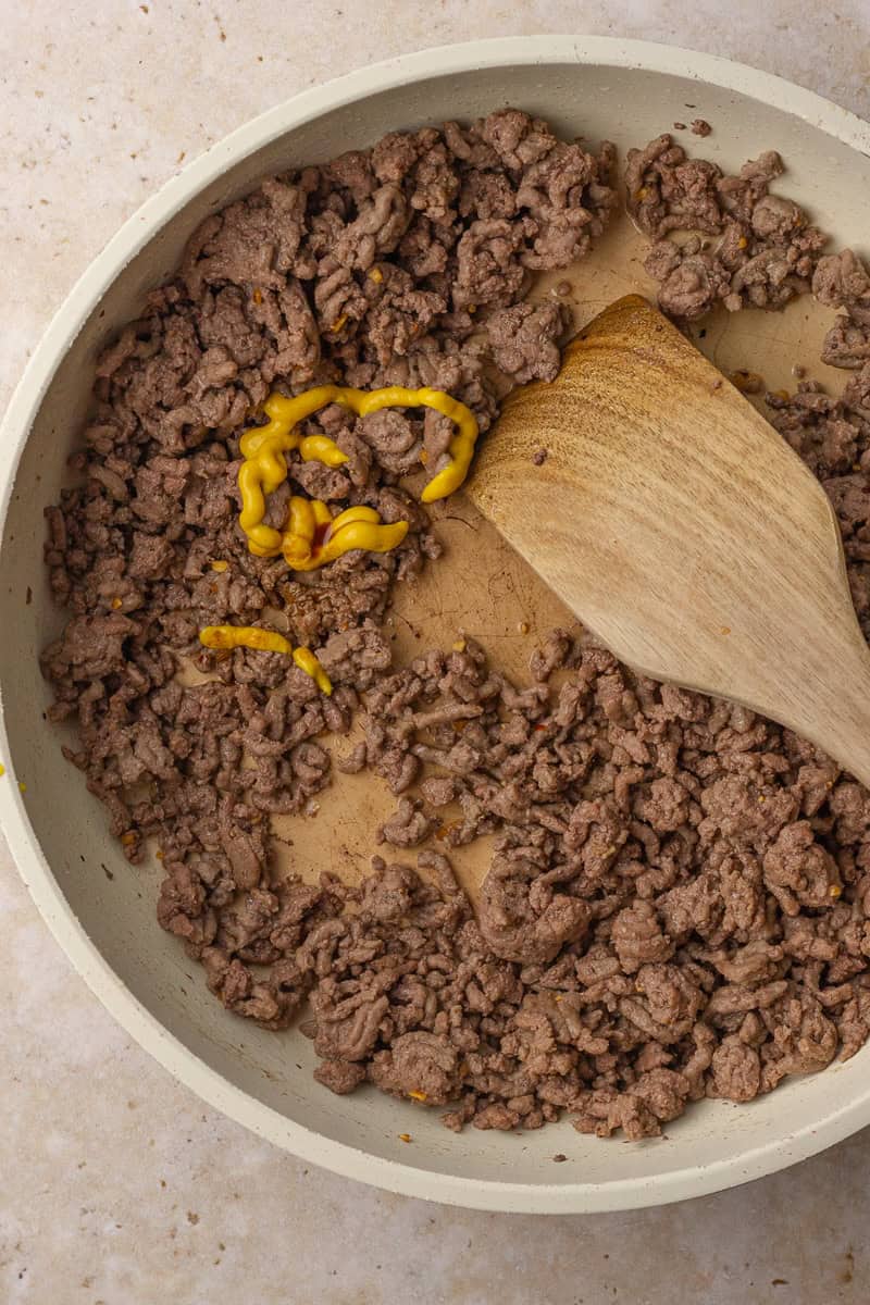 Ground beef cooking in a skillet with mustard being stirred in using a wooden spoon.