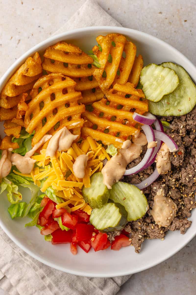 A burger bowl filled with waffle fries, beef, pickles, tomatoes, onions, cheddar, and drizzled special sauce.