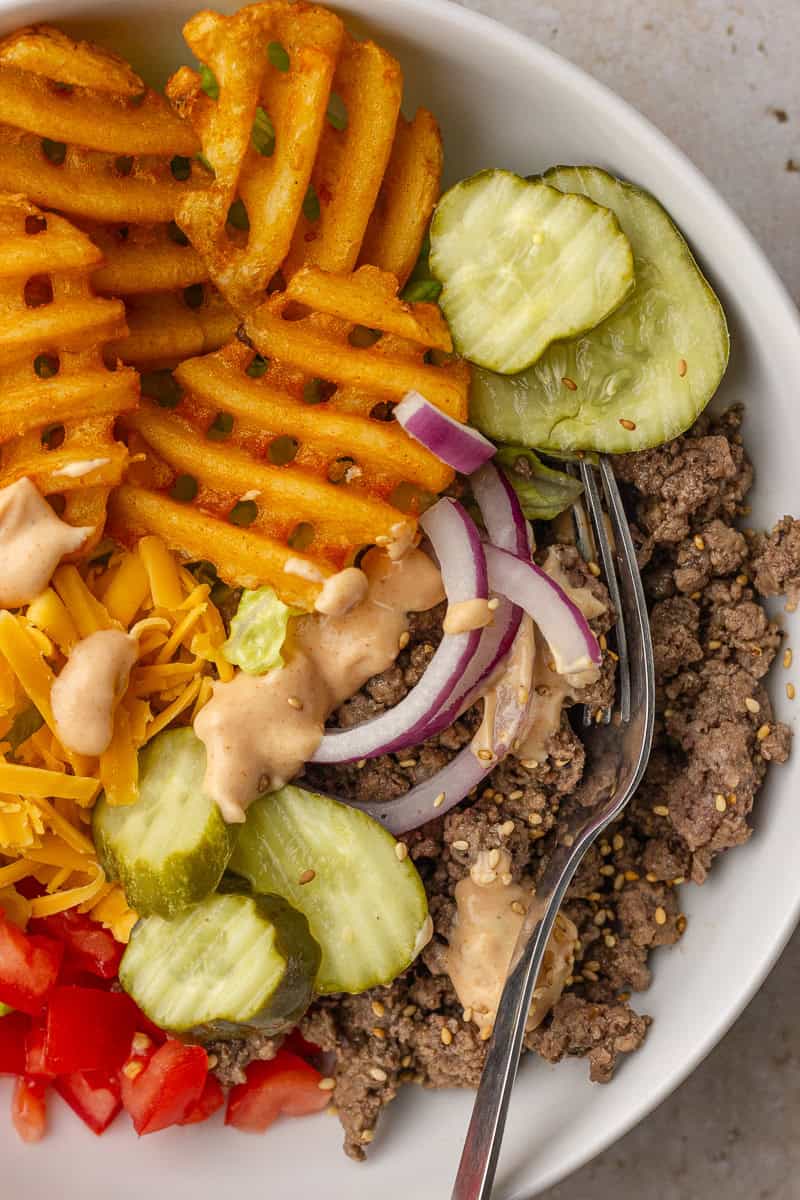 Close-up of a burger bowl with waffle fries, ground beef, cheddar, onions, pickles, tomatoes, and a fork.