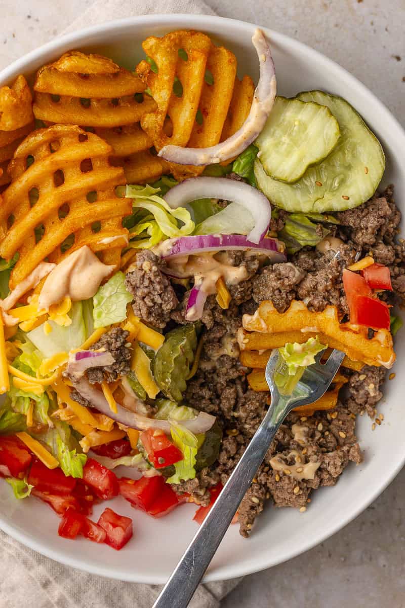 Burger bowl with waffle fries, beef, pickles, onions, cheese, tomatoes, and sauce with a fork ready to eat.