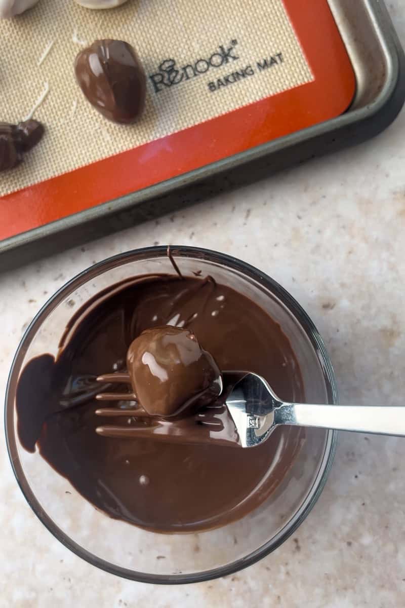 A fork lifts a dark chocolate-coated raspberry from a bowl of melted chocolate with a baking tray in the background.