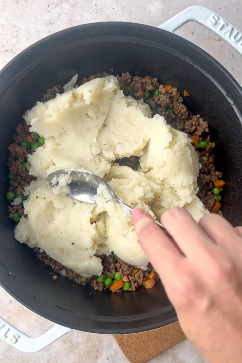Mashed potatoes being spread over beef and vegetable filling in a Dutch oven.