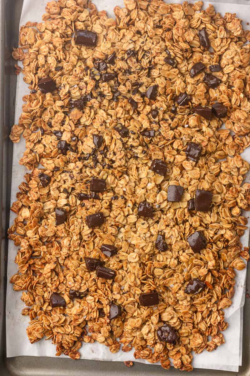 Baked granola sheet topped with chopped dark chocolate chunks and flaky sea salt cooling on parchment.
