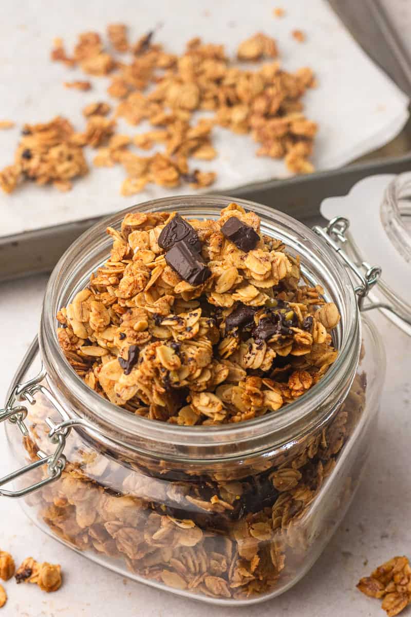 Glass jar filled with homemade granola with dark chocolate chunks and big crunchy clusters on a parchment-lined baking tray.
