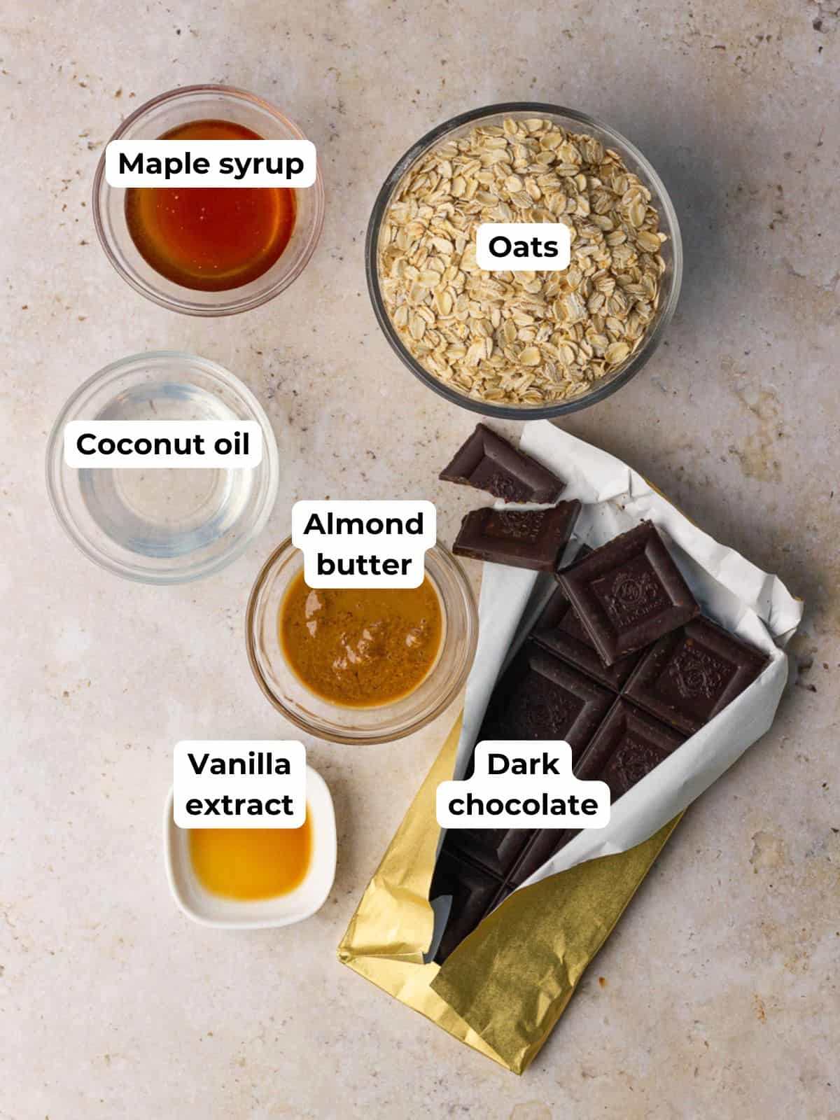 Flat lay of ingredients for granola with dark chocolate including oats, almond butter, maple syrup, coconut oil, vanilla, and a dark chocolate bar.