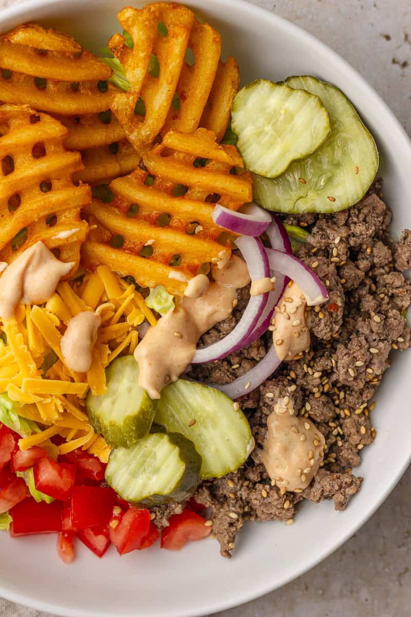 Loaded burger bowl with waffle fries, ground beef, cheddar cheese, pickles, red onion, tomato, and drizzles of healthy Big Mac sauce.