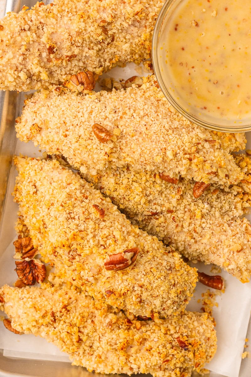 Sheet pan of pecan crusted chicken tenders with a bowl of honey mustard for dipping.