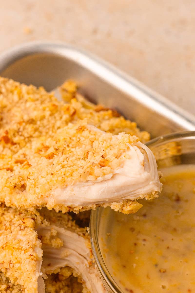 Close-up of a sliced pecan crusted chicken tender showing a juicy center beside a dipping sauce.