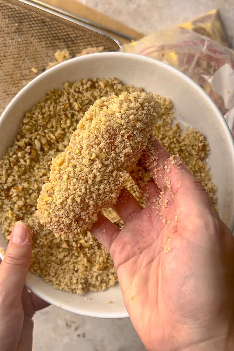 Chicken tender being pressed into a pecan panko coating in a bowl before air frying.