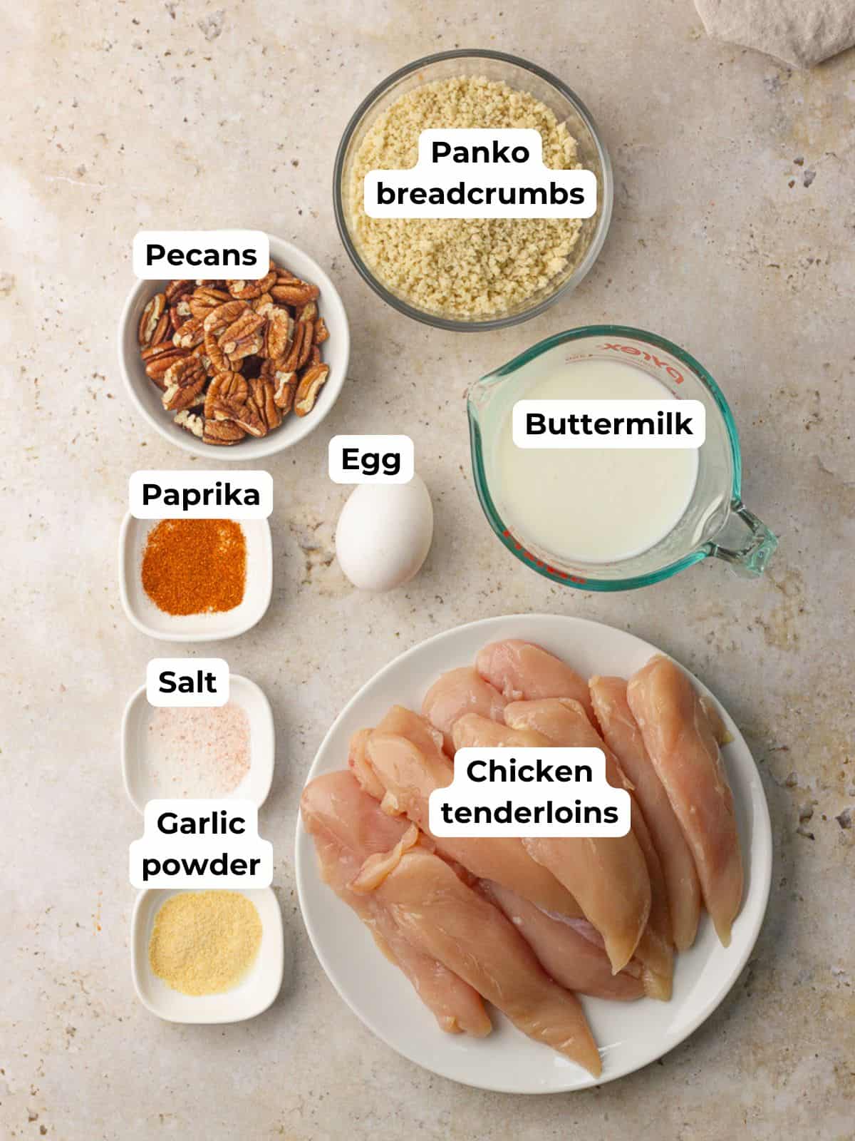 Flat lay of ingredients for pecan crusted chicken tenders including chicken tenderloins, pecans, panko, buttermilk, egg, paprika, garlic powder, and salt.