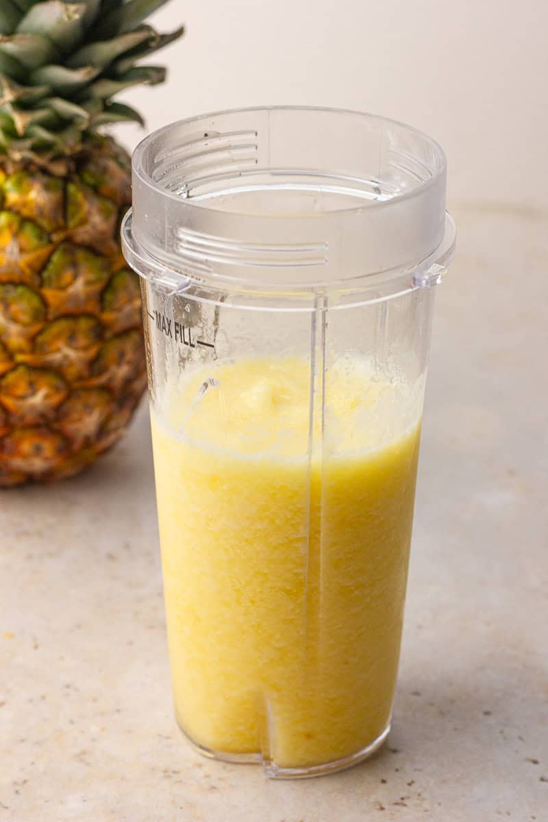 Freshly blended pineapple slushie in a blender cup with a pineapple in the background.