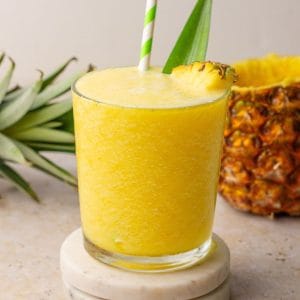 Pineapple slushie in a glass with pineapple garnish and green-striped straw.