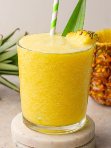 Pineapple slushie in a glass with pineapple garnish and green-striped straw.