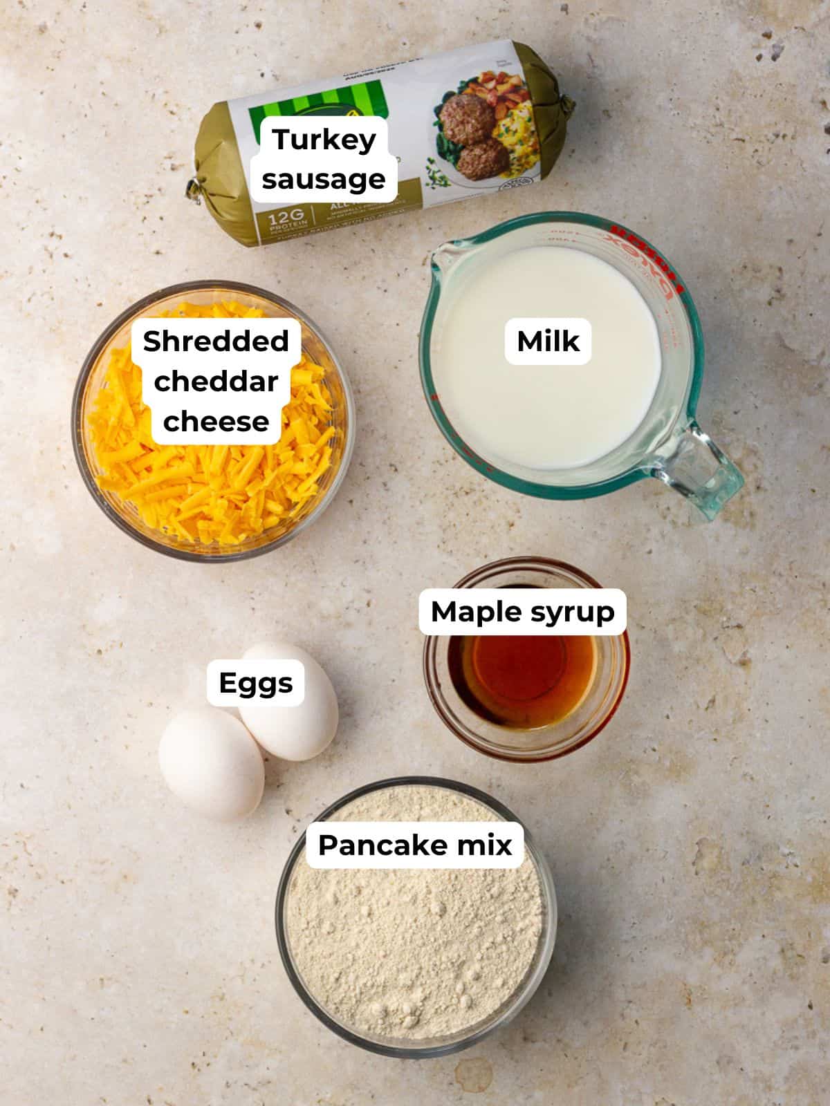 Overhead of ingredients for pancake sausage muffins including turkey sausage, pancake mix, eggs, milk, maple syrup, and shredded cheddar.