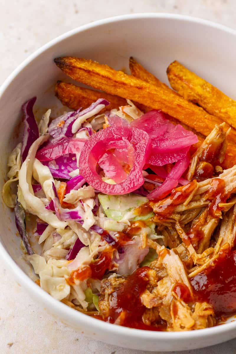 Angled view of a pulled pork bowl with fries, slaw, bright pickled onions, and barbecue sauce.
