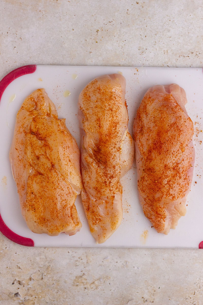 Raw chicken breasts seasoned with spices on a white cutting board.