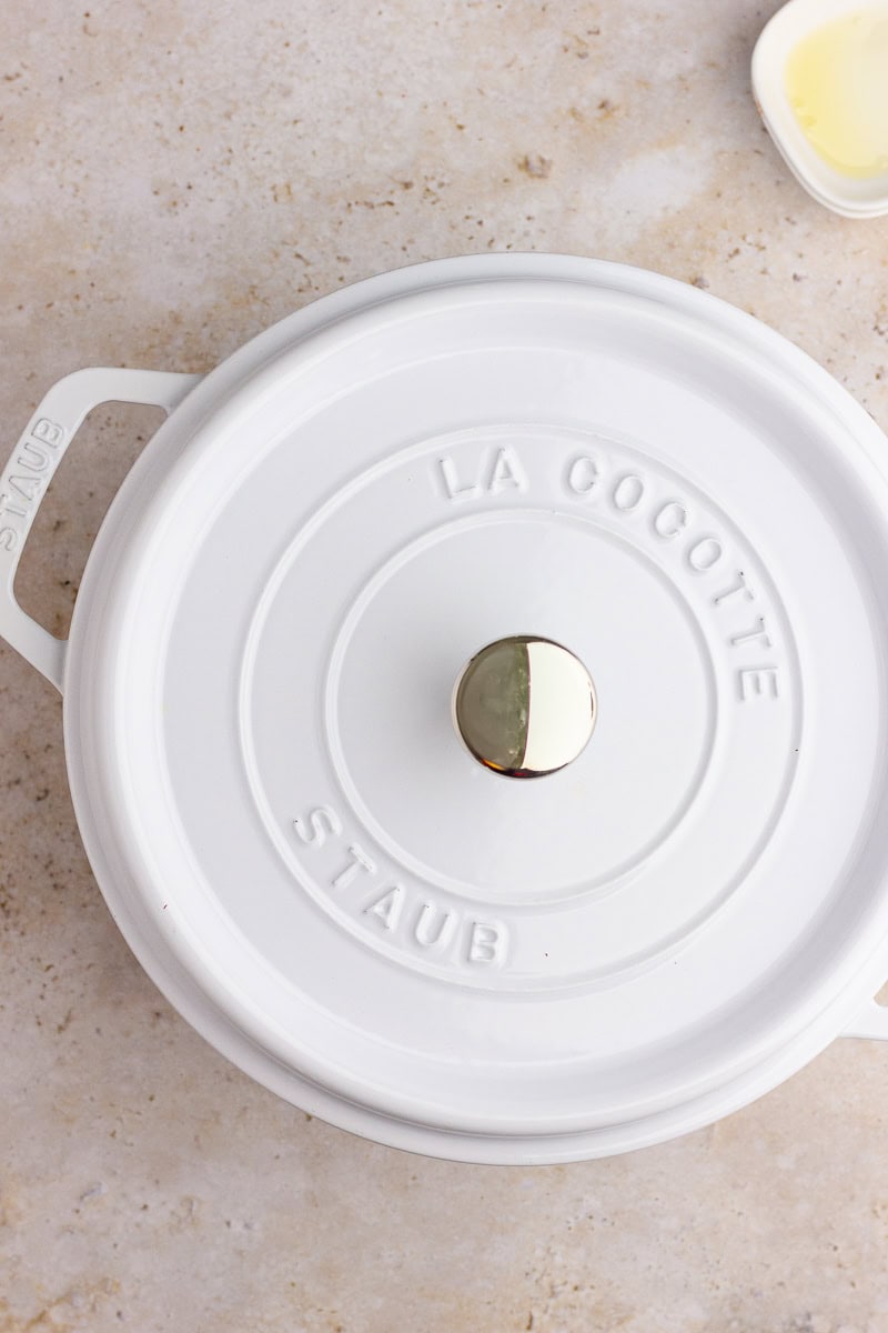 A white Staub Dutch oven with the lid on, sitting on a light stone countertop with a small dish of olive oil nearby.