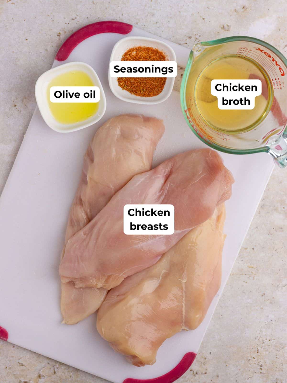 Ingredients for shredded chicken, including raw chicken breasts, olive oil, seasonings, and chicken broth labeled on a cutting board.