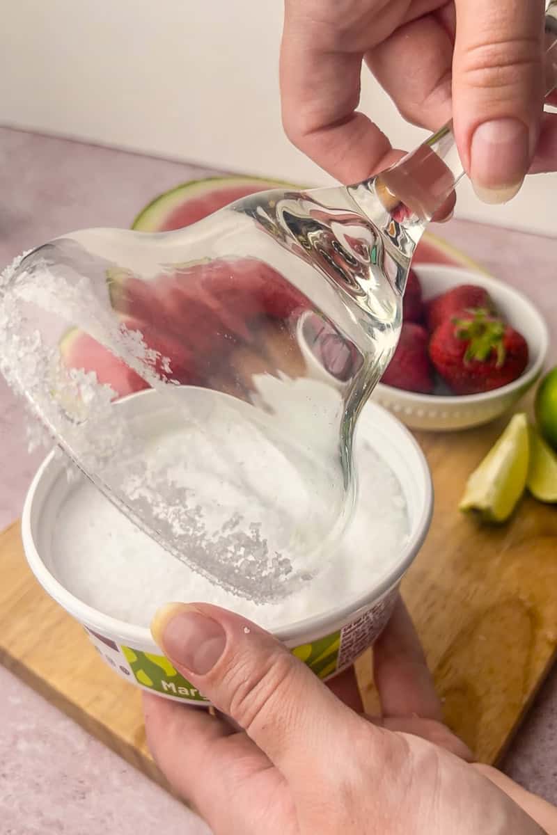 A margarita glass being dipped into coarse salt to create a salted rim, with fresh strawberries and lime wedges in the background.