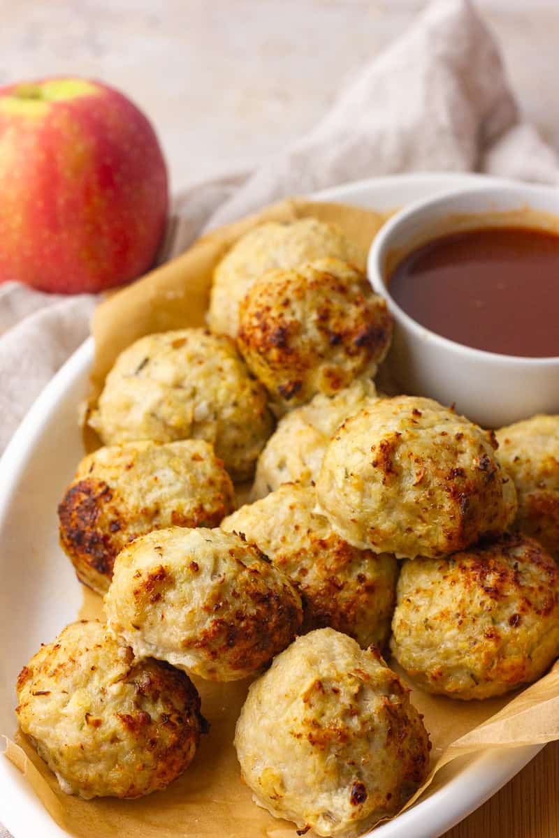 A serving dish holds golden baked apple chicken meatballs beside a small bowl of sauce and a fresh apple.