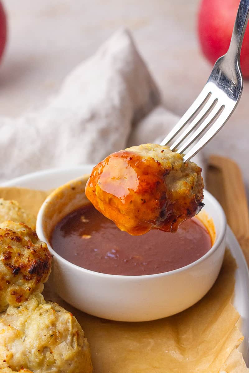 A fork lifts a glazed apple chicken meatball over a small bowl of dipping sauce.
