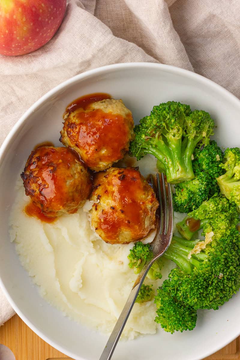 Three glazed apple chicken meatballs rest on mashed potatoes with steamed broccoli in a white bowl.