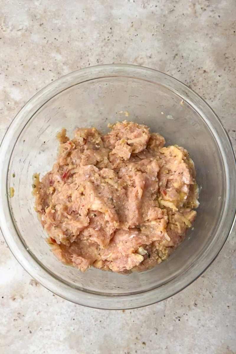 A glass bowl shows the mixed apple chicken meatball mixture with grated apple and onion.