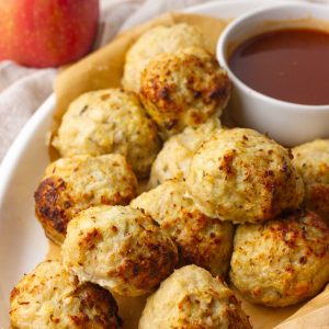 A white dish holds golden baked apple chicken meatballs beside a small bowl of sauce with a red apple in the background.