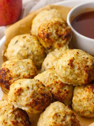A white dish holds golden baked apple chicken meatballs beside a small bowl of sauce with a red apple in the background.