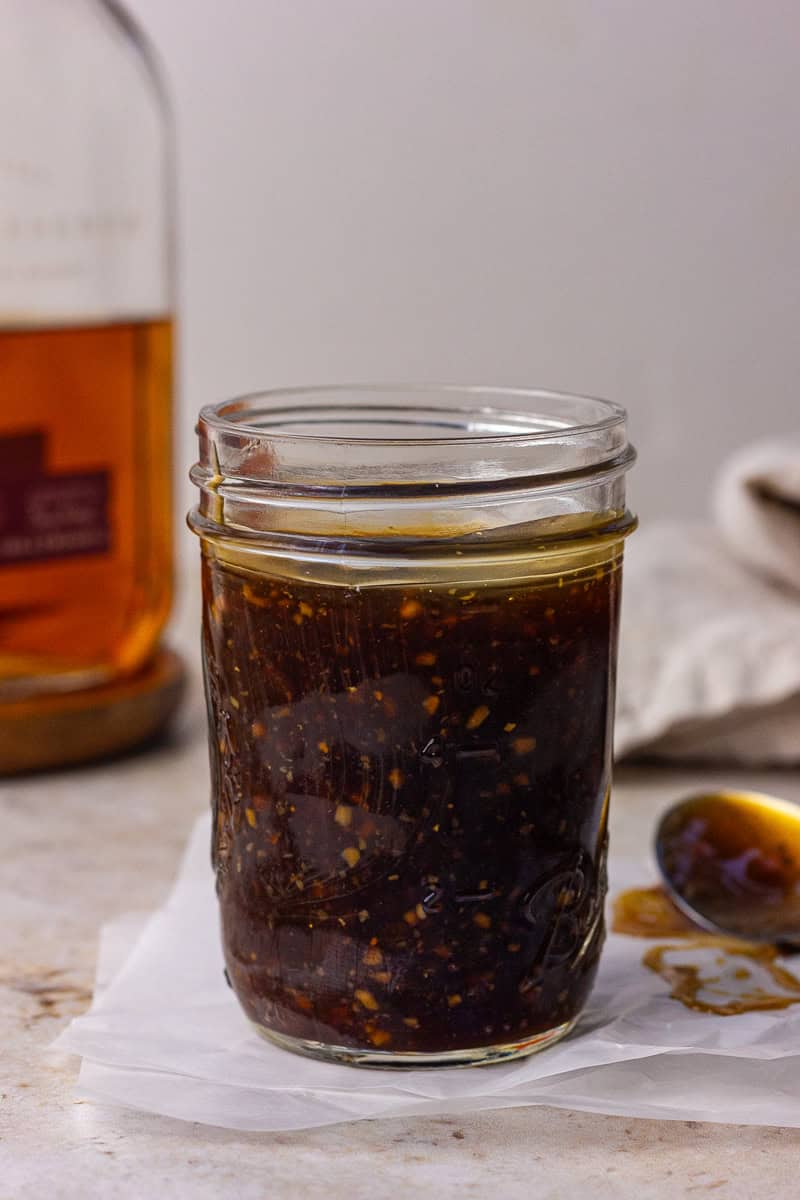 A mason jar of bourbon teriyaki sauce rests on parchment with a bourbon bottle blurred in the background and a drip-covered spoon nearby.