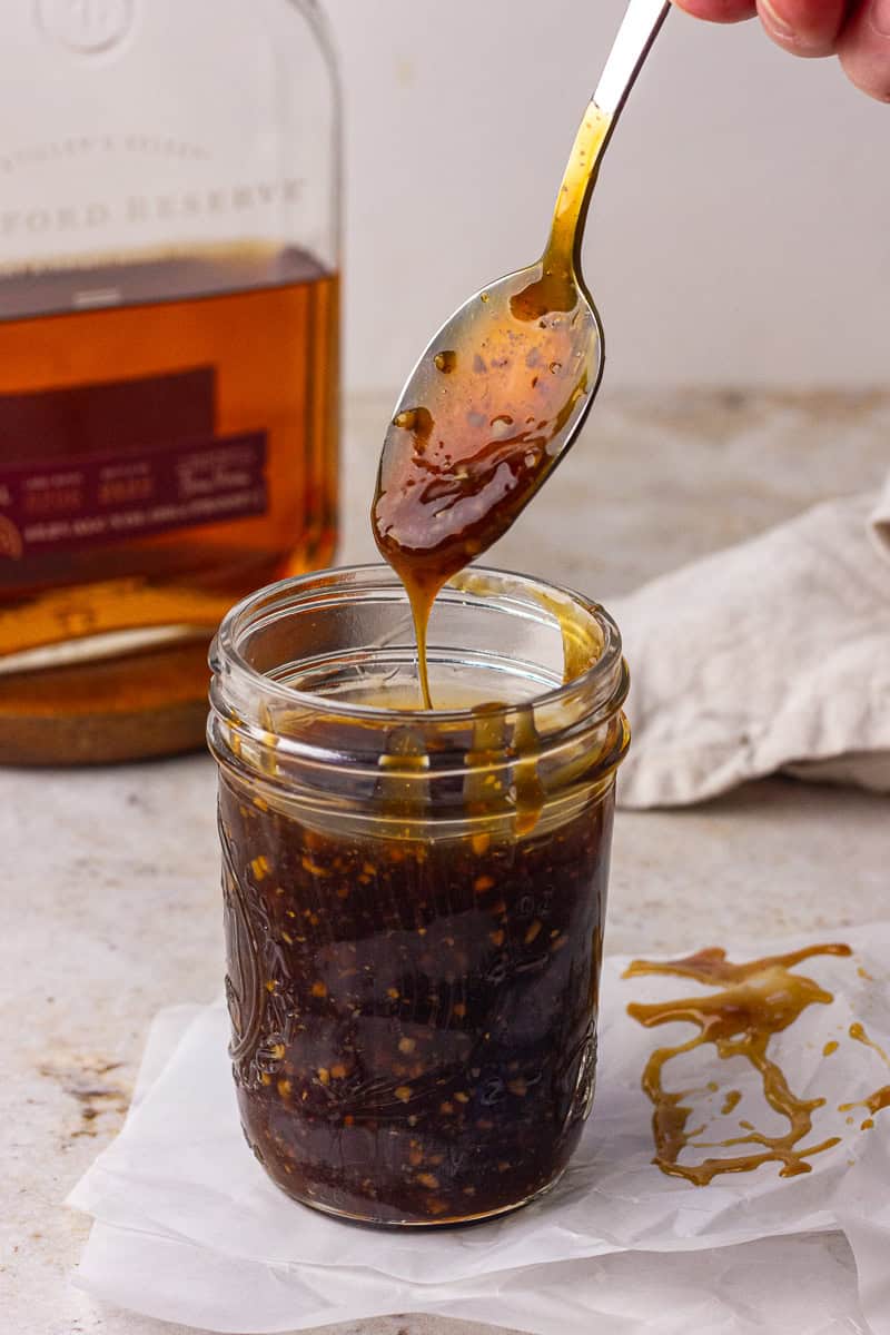 A spoon drizzles glossy bourbon teriyaki sauce into a mason jar with a bourbon bottle blurred in the background.