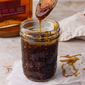 A close up shows a mason jar filled with bourbon teriyaki sauce as a spoon lifts a silky drizzle.