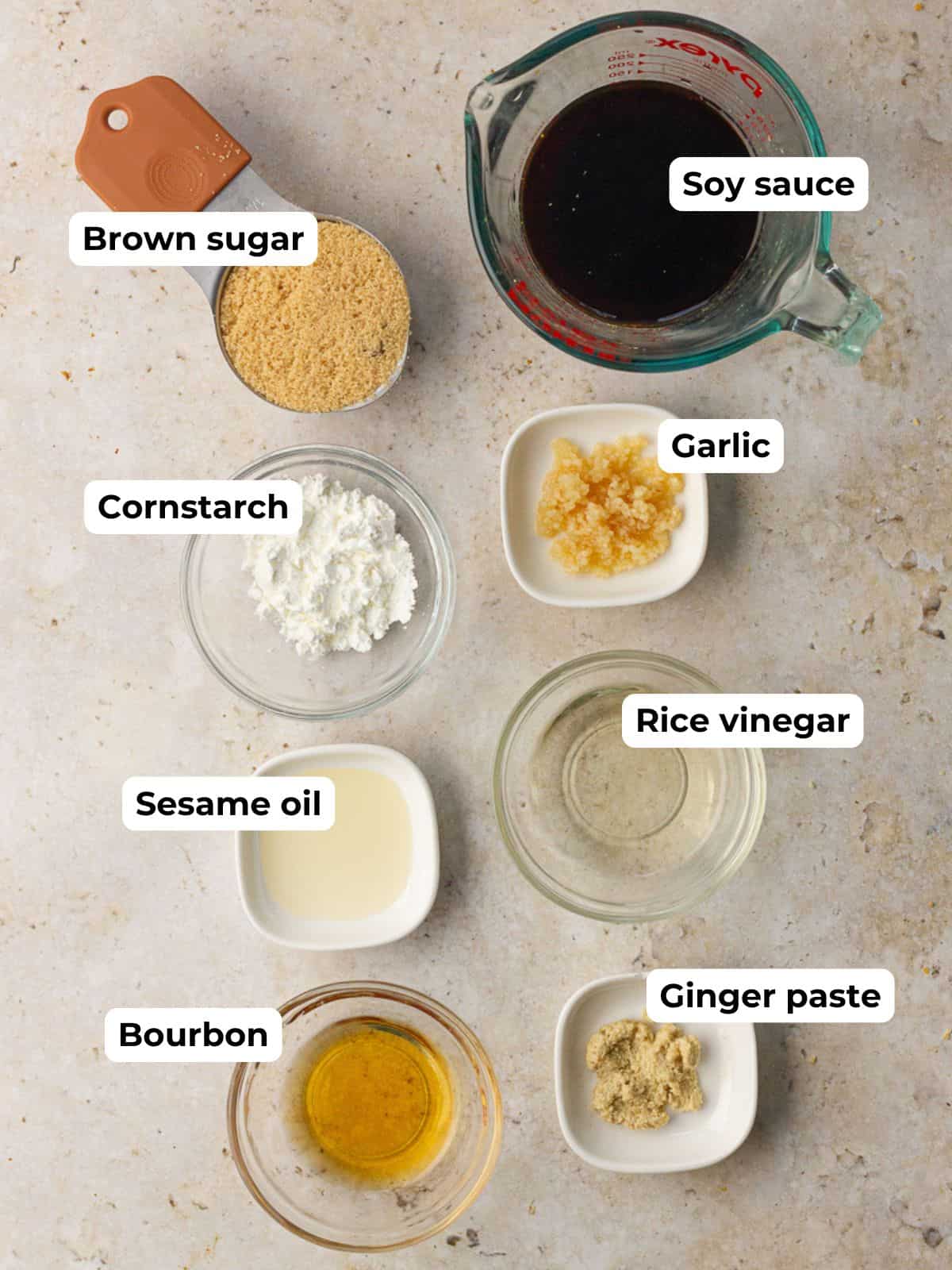 Overhead of measured ingredients for bourbon teriyaki sauce including soy sauce, brown sugar, rice vinegar, ginger paste, bourbon, garlic, sesame oil, and cornstarch.