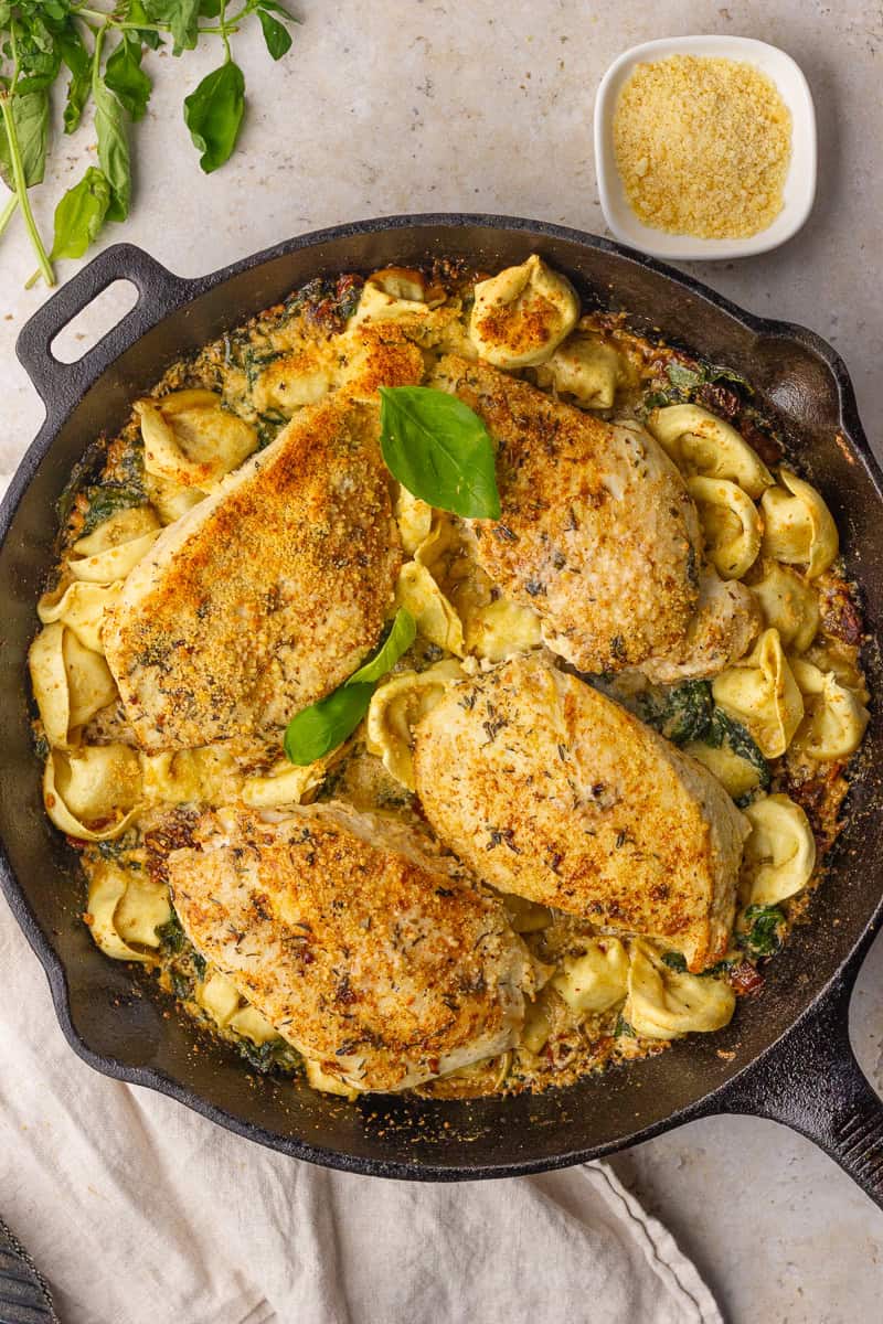 Cast-iron skillet filled with golden chicken tortellini bake topped with basil, with a small bowl of parmesan nearby.