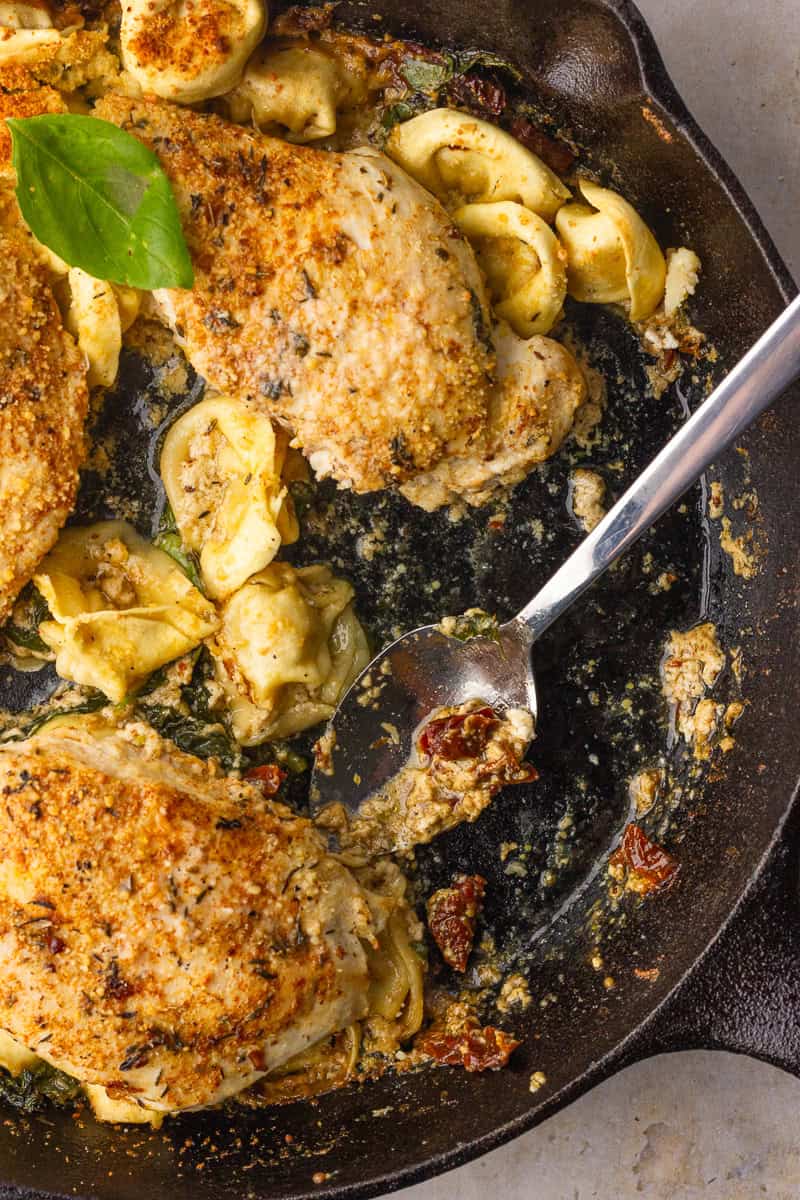 Spoon scooping creamy tortellini and sun-dried tomatoes from the cast-iron skillet beside seared chicken.